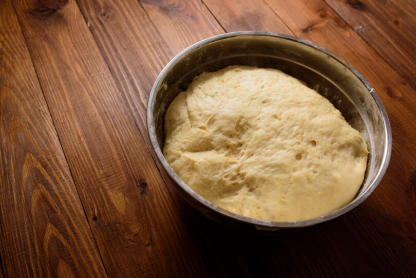 Raw yeast dough resting and rising in large metal bowl on dark wooden background. Da li znate da je kvasac pogodan za vegane i vegetarijance?